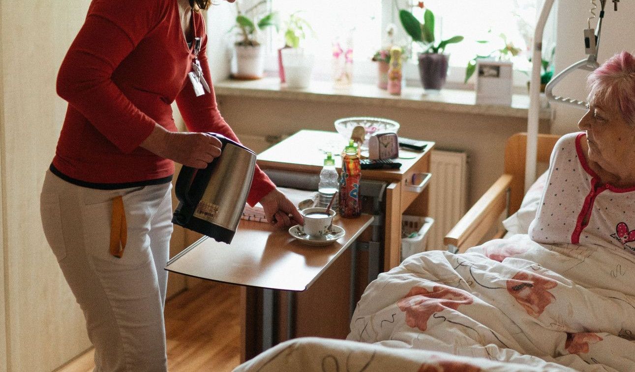 A caregiver serves tea to a senior woman in a cozy bedroom setting, fostering comfort and care.
